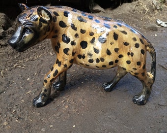Puede incluir: Una escultura de madera de una hiena con manchas negras sobre un fondo marrón. La hiena está de pie sobre sus cuatro patas con la cabeza girada hacia la izquierda. La escultura está pintada con un acabado brillante.
