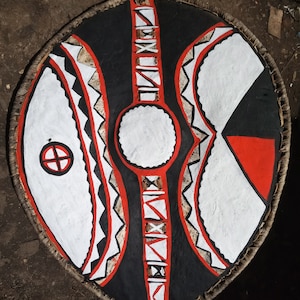 Könnte beinhalten: Ein handbemalter Schild mit einem schwarzen, weißen und roten Design. Der Schild hat eine zentrale schwarze Fläche mit weißen und roten geometrischen Mustern. Der äußere Rand ist mit gewebtem Material und einem Lederriemen versehen.