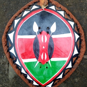 May include: A decorative shield featuring the colors of the Kenyan flag: black, red, green, and white. The shield has a brown leather border with a loop at the top for hanging. The central design includes spear-like shapes and a stylized bird.