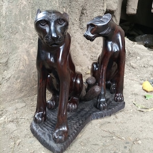 May include: Two dark brown wooden sculptures of leopards sitting on a dark brown wooden base. The leopards are facing different directions.