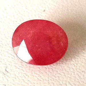 May include: A faceted, oval-shaped gemstone in a vibrant red hue. The gem has a polished surface with a prominent white facet, reflecting light. The stone is set against a neutral background, highlighting its color and shape.