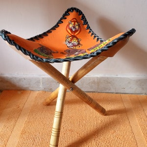 May include: A small, orange, leather folding stool with a black braided trim. The seat is decorated with a colorful print of animals, including a lion, a leopard, and a zebra. The stool has three wooden legs.