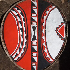 May include: A round shield with a black, white, and red geometric pattern. The shield is made of wood and has a woven rim.