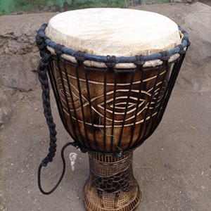 May include: A brown wooden djembe drum with a black rope handle and a white drumhead. The drum has a carved design on the side.
