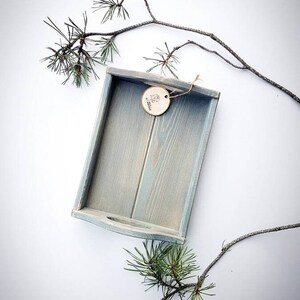 Peut inclure: Un plateau en bois bleu clair avec une étiquette ronde en bois sur laquelle est inscrit "Handmade" en lettres blanches. Le plateau est entouré de branches de pin sur un fond blanc.