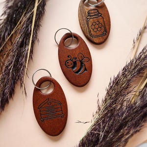 Peut inclure: Trois porte-clés en bois sur le thème de l'abeille. Les porte-clés sont de forme ovale et représentent un pot de miel, une abeille et une ruche. Les porte-clés sont en bois brun foncé et ont une finition naturelle.