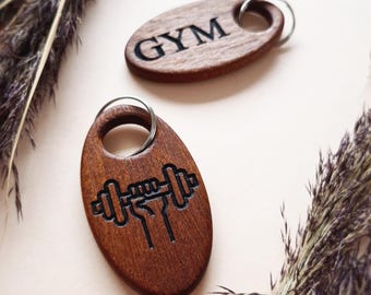May include: Two wooden keychains with a brown finish. One keychain has the word "GYM" engraved on it. The other keychain has a dumbbell with a hand holding it engraved on it.
