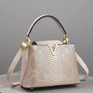 May include: Cream-colored handbag with a snakeskin pattern. The bag features a top handle, a detachable shoulder strap, and gold-tone hardware. A large V-shaped logo is on the front. The bag has a structured shape and a flap closure.