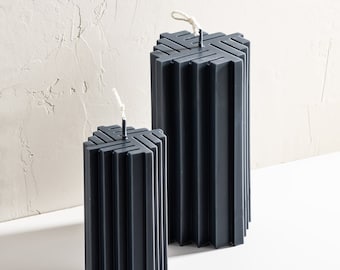 May include: Two black pillar candles with a geometric design and a white wick. The candles are on a white surface.