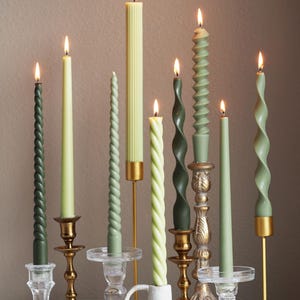 May include: A collection of lit candles in various shades of green, including spiral, ribbed, and smooth designs. The candles are displayed in ornate holders of gold, clear glass, and ceramic, creating a warm, inviting ambiance.