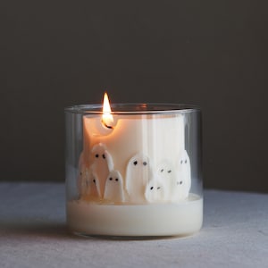 May include: A lit, white candle in a clear glass container. The candle is decorated with small, white ghost figures with black dot eyes. The flame is orange and yellow. The candle sits on a white surface.