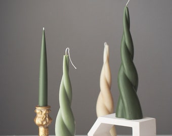 May include: An assortment of decorative candles in various shades of green and cream. The candles include a classic taper, twisted pillar candles, and a unique geometric candle holder. The candles are arranged on a white surface against a gray backdrop.