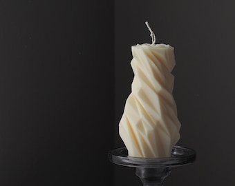 May include: A white geometric candle sits on a grey glass candle holder. The candle is tall and has a textured, faceted design.