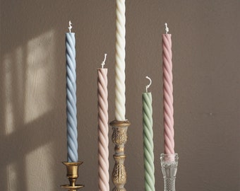 May include: Five twisted candles in different colors, including light blue, pink, cream, and green, are displayed on various candle holders. The holders are made of gold metal and clear glass.
