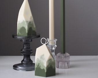 May include: Three candles in different shapes and sizes. A tall, thin white candle, a shorter, thicker green candle, and a geometric green and white candle on a black pedestal.