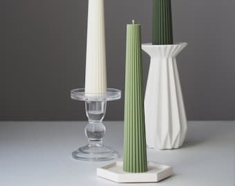 May include: Three tall candles on different holders. The candles are ribbed and have a white, green, and dark green color. The holders are a clear glass candlestick, a white ceramic base, and a white ceramic base with a geometric design.