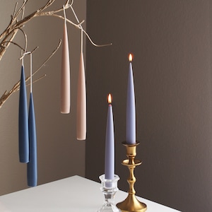 May include: A set of taper candles in various colors, including pink, blue, and gray. The candles are arranged on a white table with a gold candlestick and a clear glass candle holder.