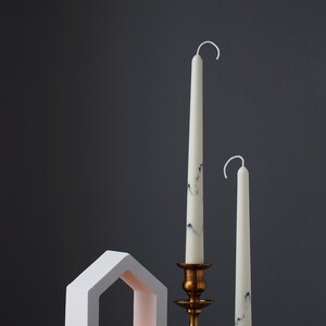 May include: Two white candles, one with black marbling, on a gold candlestick and a white house-shaped candle holder with a lit tea light inside. The candles are on a wooden surface against a dark gray background.