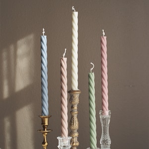 May include: Five twisted candles in different colors, including light blue, pink, cream, and green, are displayed on various candle holders. The holders are made of gold metal and clear glass.