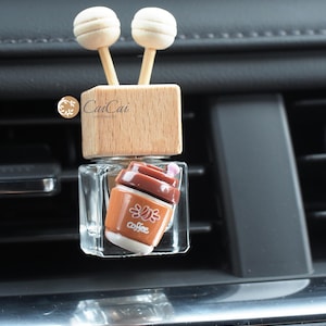 May include: A car air freshener with a wooden base and two wooden sticks. The bottle is clear glass and contains a brown liquid. The bottle has a small, brown coffee cup charm attached to it. The charm has the word "Coffee" printed on it.
