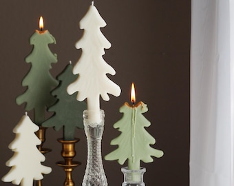 Peut inclure: Plusieurs bougies en forme de sapin de Noël, blanches et vertes, dont certaines sont allumées. Les bougies sont présentées sur différents bougeoirs anciens, en verre transparent et en métal doré. La scène se déroule sur une surface blanche.