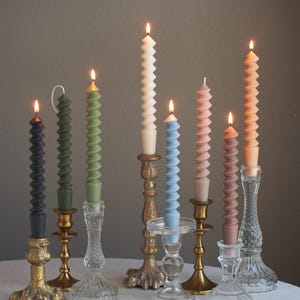 May include: Seven candlesticks with lit candles in various colors, including black, green, white, blue, and pink. The candles are all spiral shaped and are on a white tablecloth.