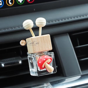 May include: A small glass bottle containing a red and white mushroom charm. The bottle is attached to a wooden block with two wooden sticks topped with white balls. The bottle is labelled "CaiCai Handmade".