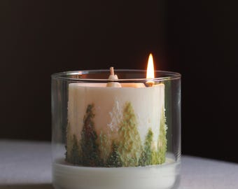 May include: A lit, white candle in a clear glass holder. The candle features green tree designs. The flame is bright orange. The candle sits on a white surface against a dark background.