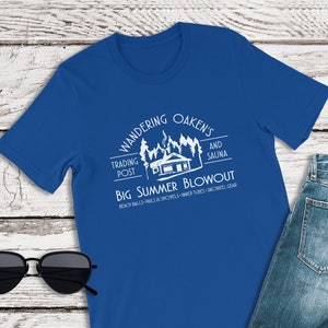 Puede incluir: Una camiseta azul real con un diseño gráfico blanco de una cabaña con una chimenea y humo saliendo de ella. El texto en la camiseta dice "Wandering Oaken's Trading Post and Sauna Big Summer Blowout Beach Balls - Balls & Shovels - Tires - Snorkel Gear".