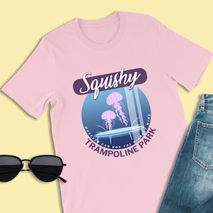 May include: Pink t-shirt featuring a graphic of a trampoline with jellyfish jumping on it. The text on the graphic reads "Squishy Trampoline Park Who can hop the highest wins".