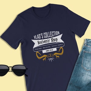 May include: A navy blue t-shirt with a white graphic design that says "Vlad's Collection Souvenir Shop" with a gold ribbon and unicorn, duckling, and little things written below. The text "Since 2010" is also included in the design.