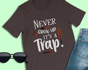 May include: A dark brown t-shirt with the text "NEVER GROW UP IT'S A TRAP!" in white. The text is surrounded by small brown and orange illustrations of tools and feathers.