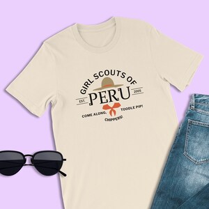 May include: A beige t-shirt with a graphic design featuring a brown hat with a red bow and the text "Girl Scouts of Peru Est. 2005 Come Along. Toodle Pip! Chippers!"