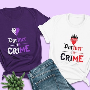 May include: Two purple and white t-shirts with the text "Partner in Crime" and a heart design. The purple shirt has a white heart with a purple swirl design. The white shirt has a red heart with a crown on top. The shirts are laid out on a light purple background with a pair of blue jeans and a small green plant.
