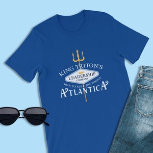 May include: A royal blue t-shirt with a gold graphic of a trident and a sign that reads "King Triton's Leadership Company How to Rule the Whole Atlantica".