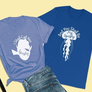 May include: Two t-shirts in blue and light blue colours. The blue shirt features a white jellyfish silhouette with the text "And You Shall Be My Squishy". The light blue shirt has a white silhouette of a jellyfish with the text "I Shall Call You Squishy".