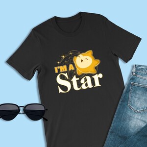 May include: Black t-shirt with a yellow cartoon star with the text "I'm a Star" on it. The t-shirt is on a blue background with a pair of sunglasses and blue jeans.