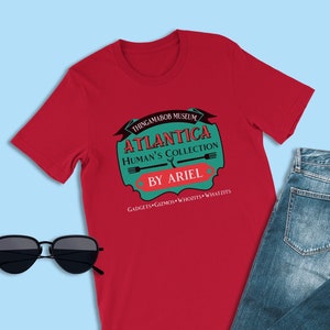 Può includere: Maglietta rossa con un logo verde e bianco che dice "Thingamabob Museum Atlantica Human's Collection By Ariel Gadgets Gizmos Whoozits Whatzits"