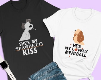 Puede incluir: Dos camisetas gráficas, una negra y otra blanca, con ilustraciones de perros y texto. La camiseta negra dice "She's my Spaghetti Kiss", y la camiseta blanca dice "He's my Lovely Meatball". Un par de vaqueros azules y un smartwatch blanco también están en la imagen.
