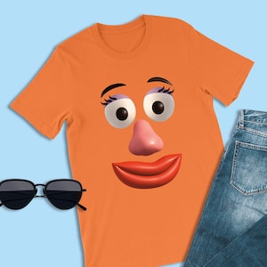May include: Orange t-shirt featuring a cartoon face with large eyes, a pink nose, and red lips. The shirt is paired with blue jeans and black sunglasses. The image is set against a light blue background.