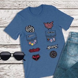 May include: A blue t-shirt with various patches, including a dragon, a sword, a crown, and the words "SMOTE" and "CREBBID". The t-shirt is paired with blue jeans.