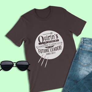 Może przedstawiać: Brązowy t-shirt z białym kołem z napisem "Quirrin's Academy Leadership Curriculum Be the Future Leader! Since 2017".