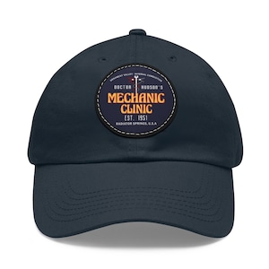 Puede incluir: Una gorra de béisbol azul marino con un parche cosido que dice "Doctor Hudson's Mechanic Clinic Est. 1951 Radiator Springs, U.S.A."