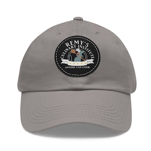 Può includere: Un cappello da baseball grigio con una toppa bianca e nera che presenta un cuoco topo dei cartoni animati che tiene un cucchiaio di legno. La toppa recita "Remy's Culinary Institute Est. 2007 Anyone Can Cook".