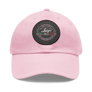 Könnte beinhalten: Hellrosa Baseballkappe mit gebogenem Schirm. Auf der Kappe befindet sich ein kreisförmiger Aufnäher mit dem Text "Luigis" und "Casa Della Tire" in einem Vintage-Design. Der Aufnäher enthält grüne, weiße und rote Streifen und den Text "Get 2 Snow Tires For Free".