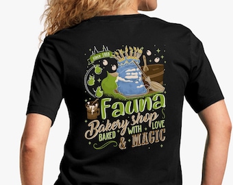Op de afbeelding: Zwart T-shirt met een afbeelding van een blauwe ketel met groene appels, een bezem en de tekst "Fauna Bakery Shop Baked with Love & Magic" in bruin en witte letters.