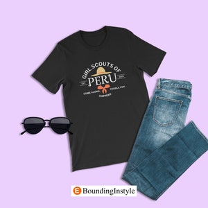 May include: Black t-shirt with a white graphic of a girl scout patch. The patch features a straw hat with a red bow and the text "Girl Scouts of Peru Est. 2005 Come Along. Toodle Pip! Chippers!"