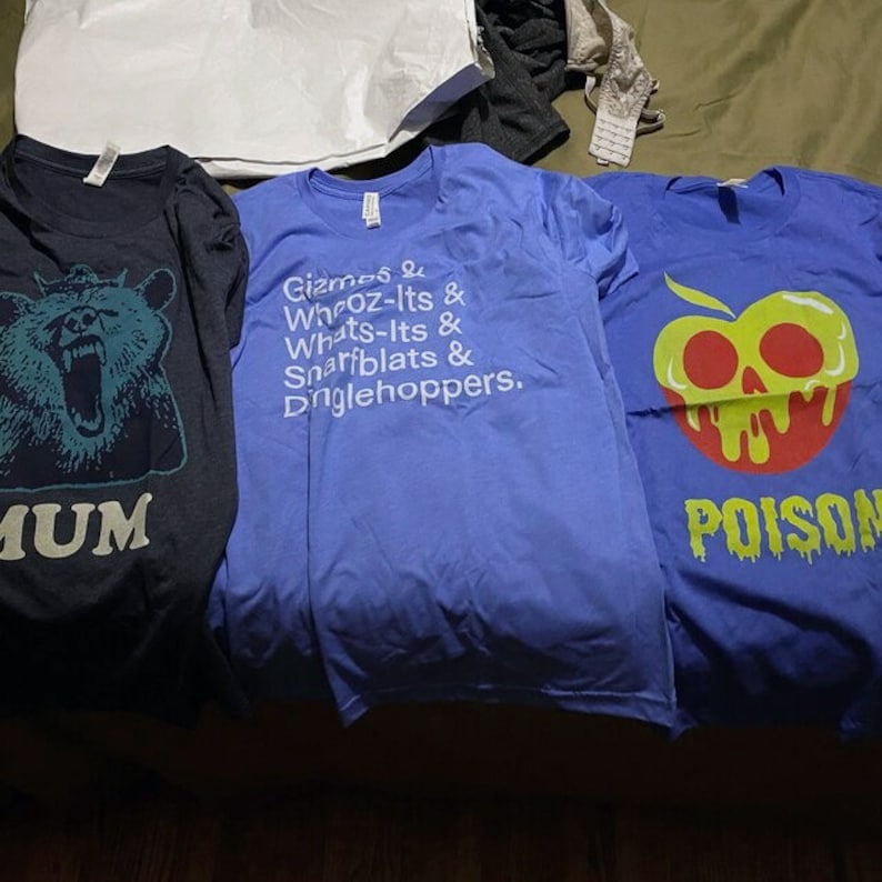 May include: Three t-shirts with different designs. The first is dark blue with a bear and the word "MUM". The second is light blue with white text that reads "Gizmos & Whooz-Its & Whats-Its & Sparfblats & Dinglehoppers.". The third is blue with a red apple with a skull and bones and the word "POISON" in yellow.