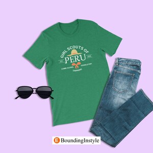 May include: Green t-shirt with a white graphic of a Girl Scouts of Peru logo. The logo features a straw hat and the text "Girl Scouts of Peru" with the text "Est. 2005" and "Come Along, Toodle Pip! Chippers!" underneath.