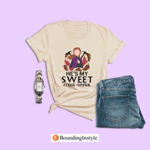 May include: A beige t-shirt with a graphic of a cartoon character with brown hair and a purple dress. The text "He's my sweet fixer-upper" is printed below the graphic.
